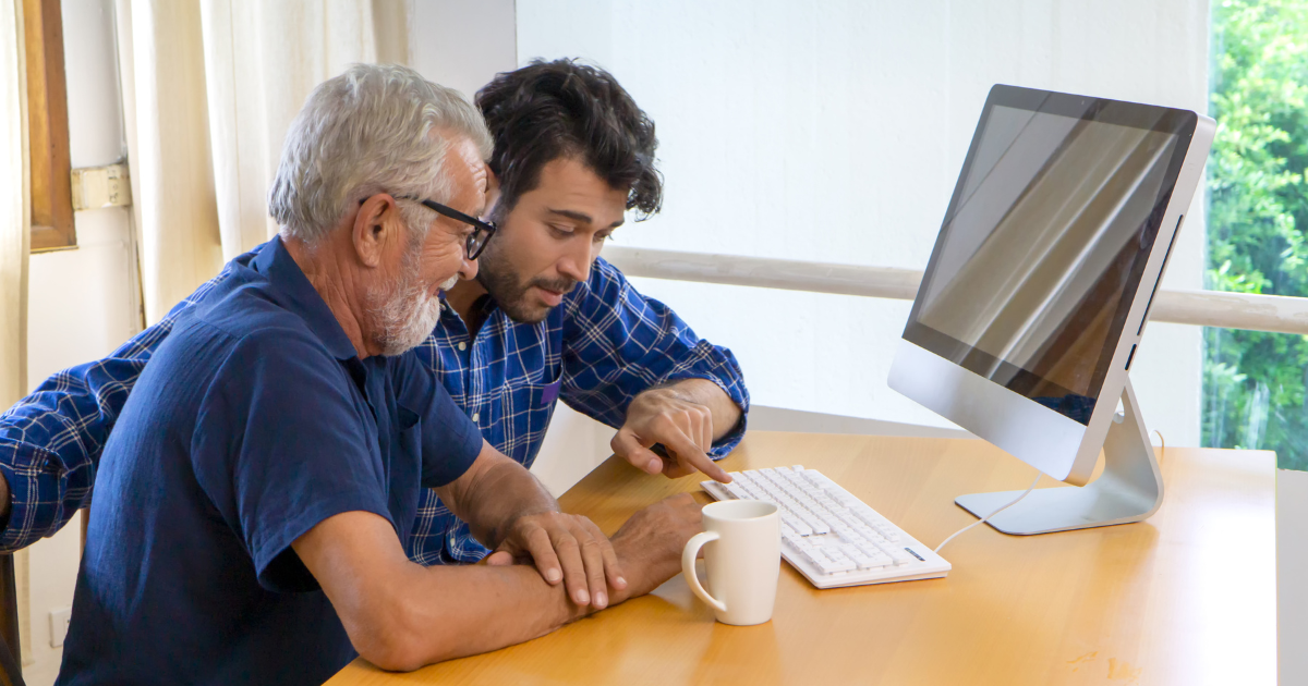 A son who is caregiving for an elderly father helps him with his computer.