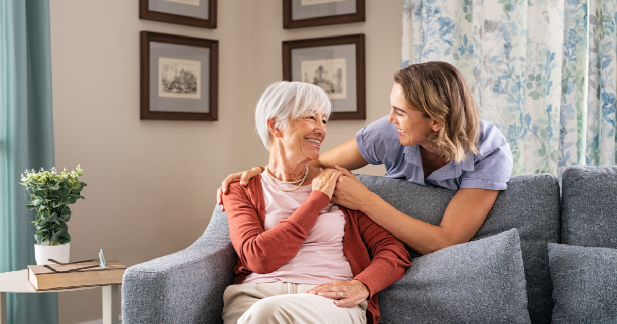 Smiling caregiver with hand over the shoulder of a happy elderly woman sitting on the couch, representing how home care services can help the recipient of care and caregiver stay happy and healthy.