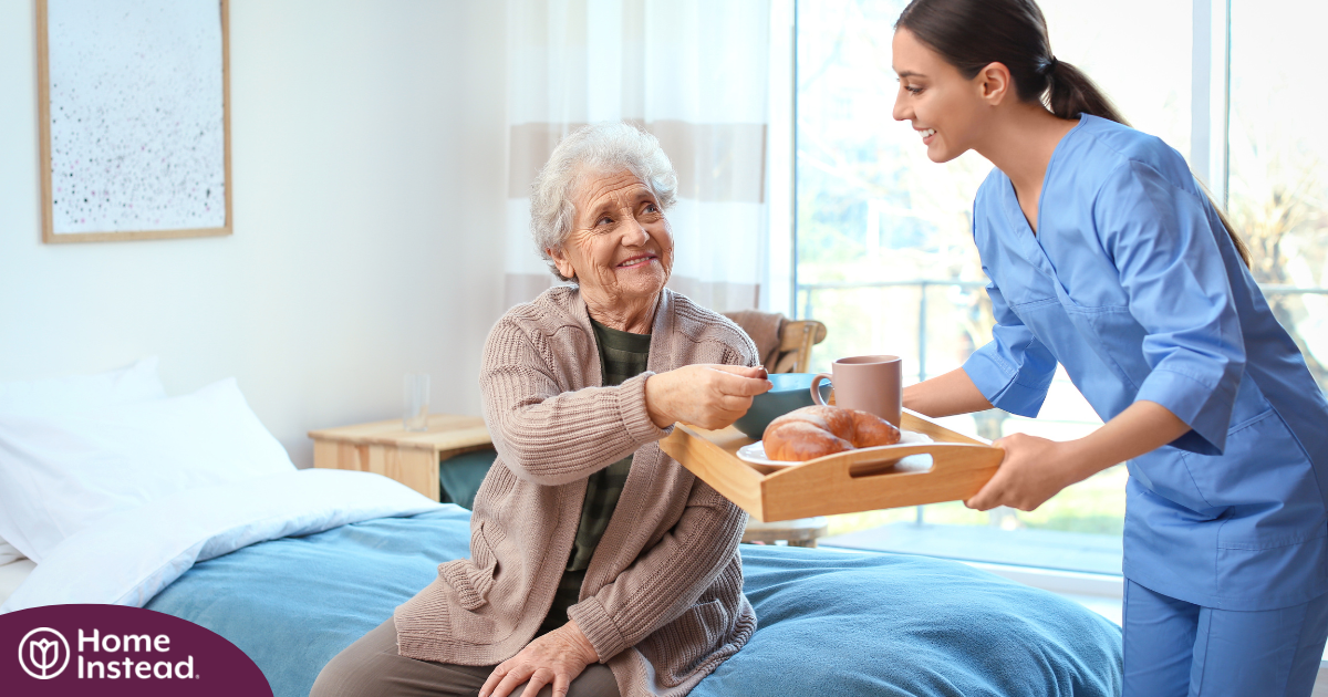 A professional caregiver smiles and provides food to an older adult in her care, representing the compassionate care needed for those with dementia.